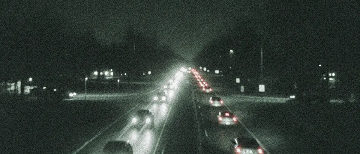 Night view from a traffic camera: headlight beams and tail lights on a dark road, vehicle outlines barely visible